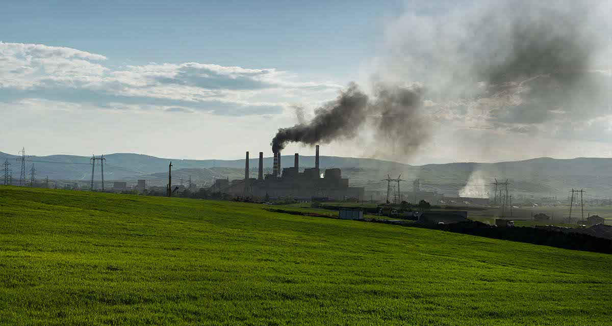 Fumi neri si alzano dalle ciminiere di un impianto industriale in un paesaggio di campagna.