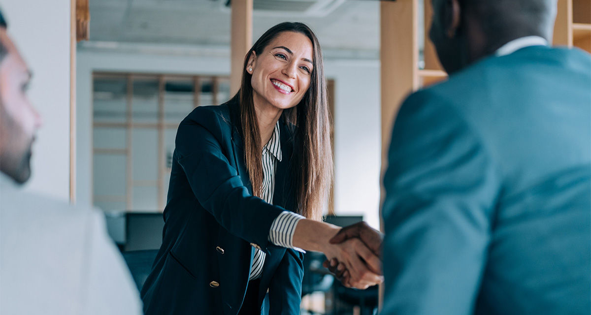 Una professionista accoglie sorridente e in piedi un cliente, stringendogli la mano.