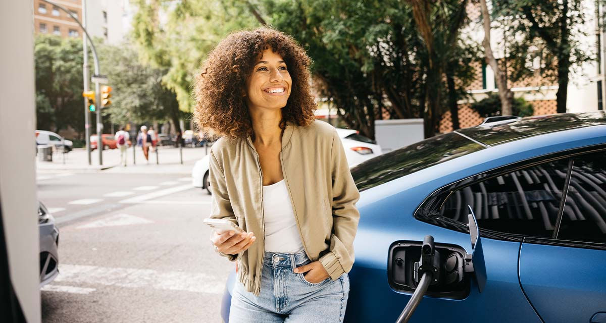 Una ragazza sorride e consulta lo smartphone mentre la sua auto elettrica è in ricarica
